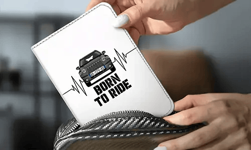 Person holder hvid pung med sort bil og hjerteslagsdesign, BORN TO RIDE tekst, kulfiber-taske i baggrunden