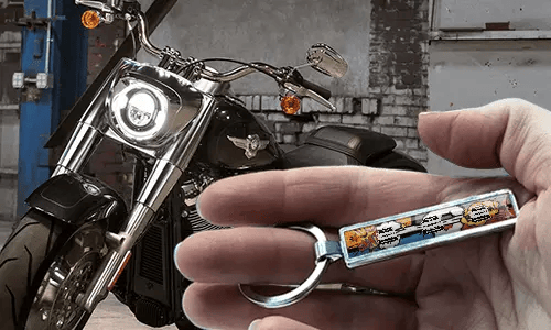 Comic-style license plate keychain held in a hand with a motorcycle in the background