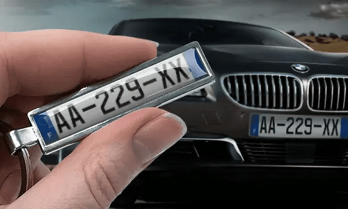 Comic-style license plate keychain held in a hand in front of a car with matching plates